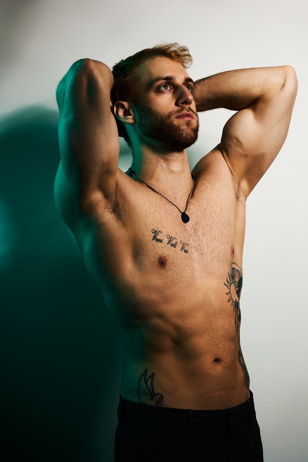 Shirtless athletic model with tattoos poses dramatically against turquoise wall with dramatic lighting and shadows.