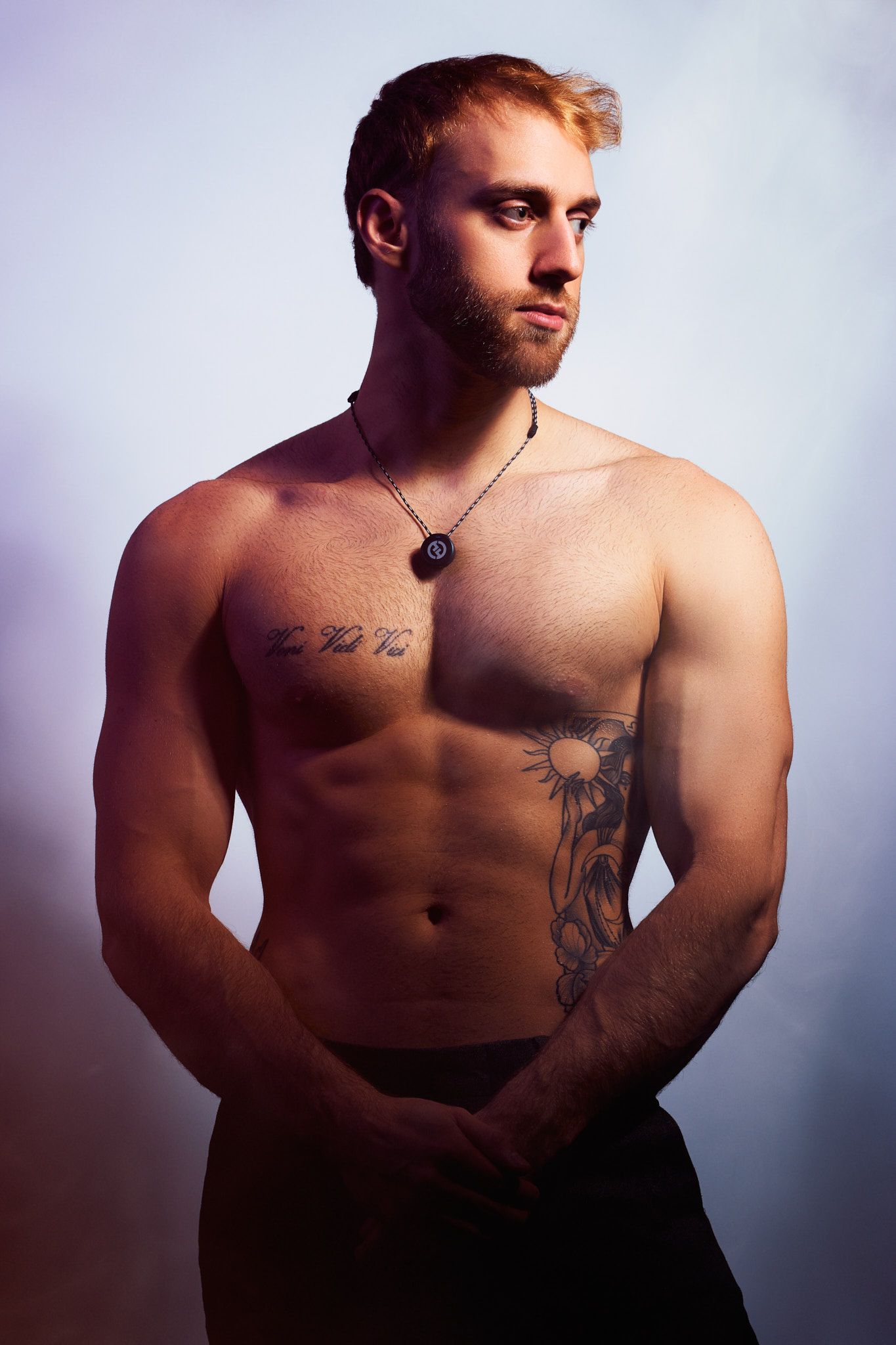 A shirtless person with tattoos poses against a dark background in dramatic lighting.
