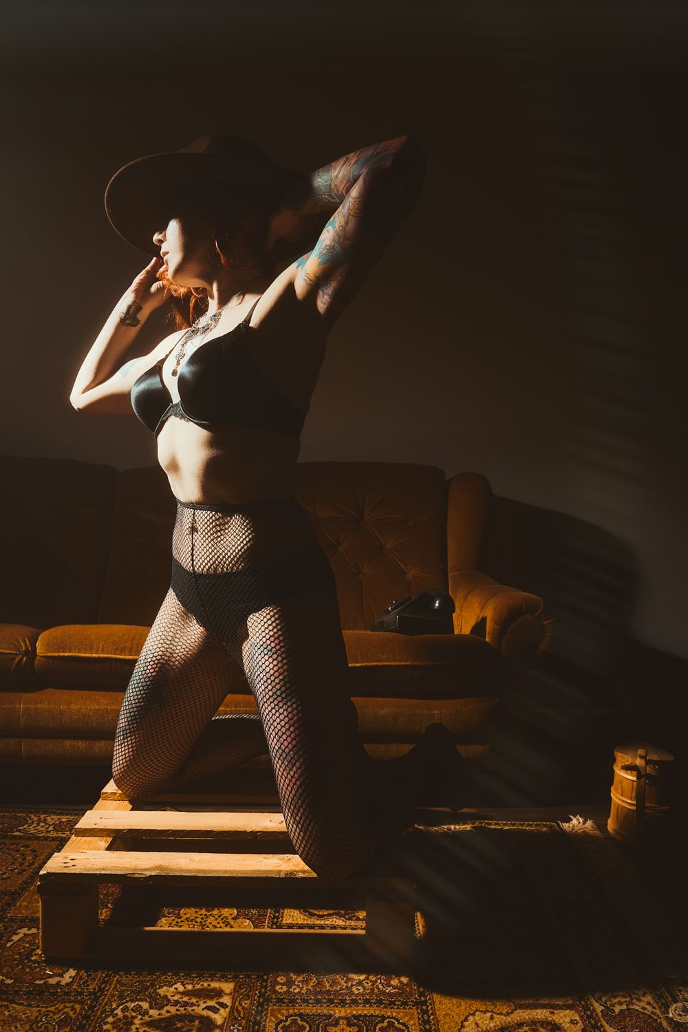 Woman in black lingerie, fishnet tights, and wide-brim hat posing on wooden pallet in dramatic moody lighting.