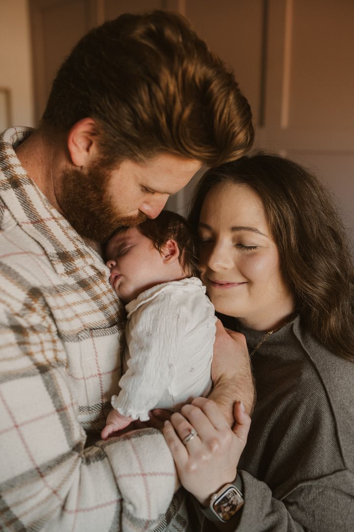A family shares an intimate moment as they embrace their newborn baby in warm, moody lighting.
