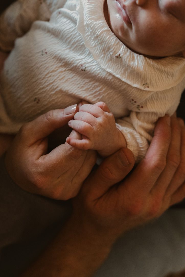 A small baby in white clothing grasps their hands together while being cradled in adult hands.