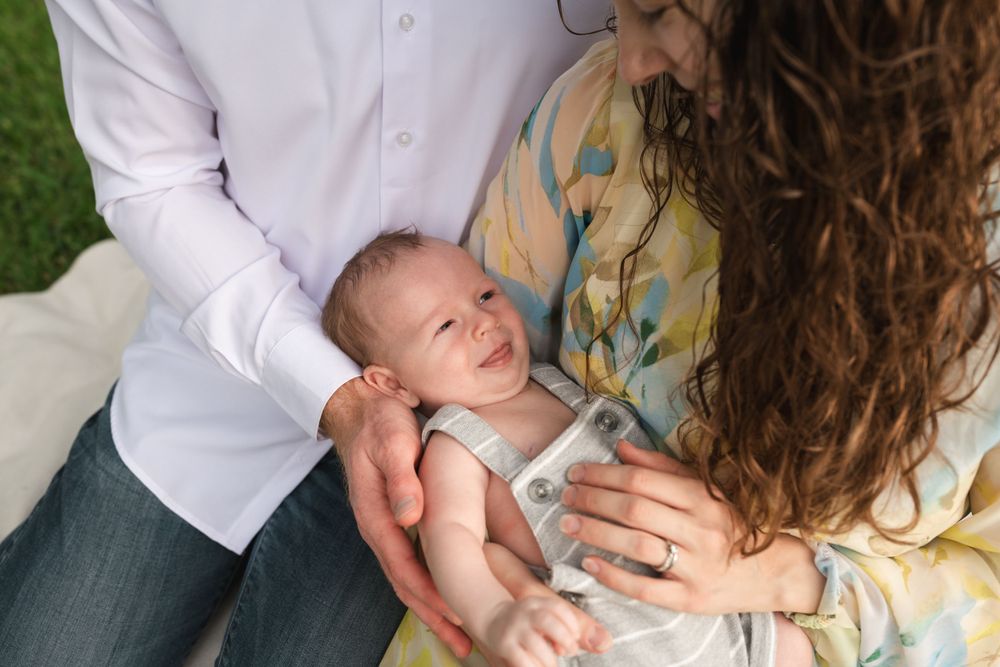 Baby in grey overalls smiling during outdoor family photoshoot.