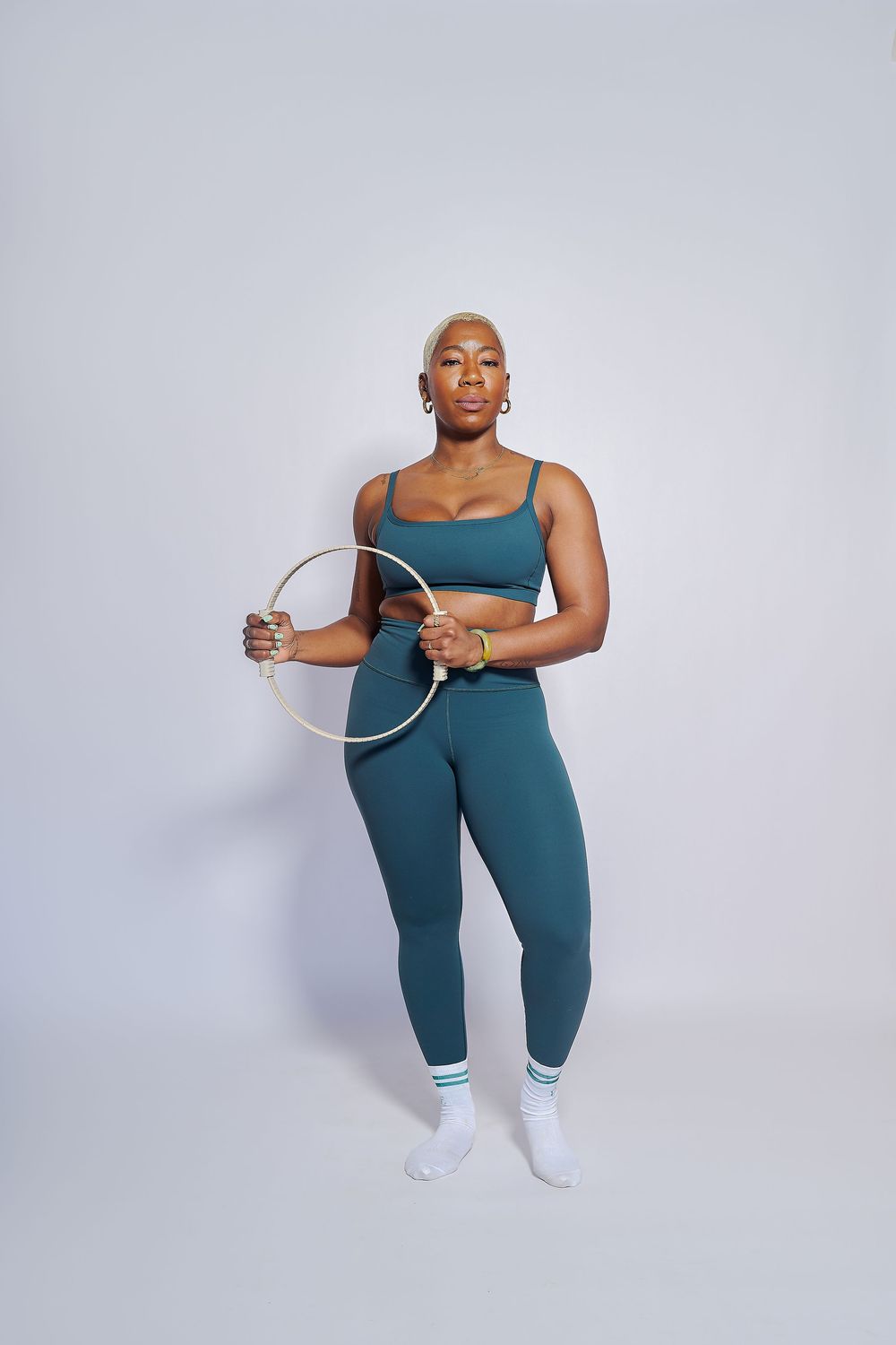 Athletic Black woman with blonde hair in teal sports set holding a hula hoop in a studio in the Bay Area.