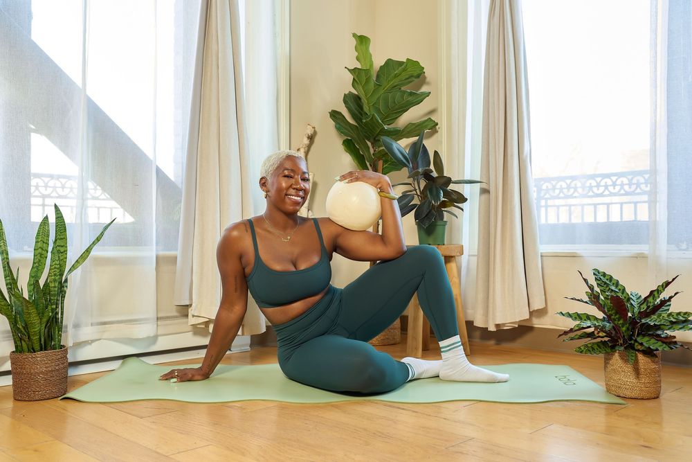 Smiling Black woman in athletic wear doing yoga on a mat surrounded by houseplants for a branding shoot in a Bay Area photography studio.