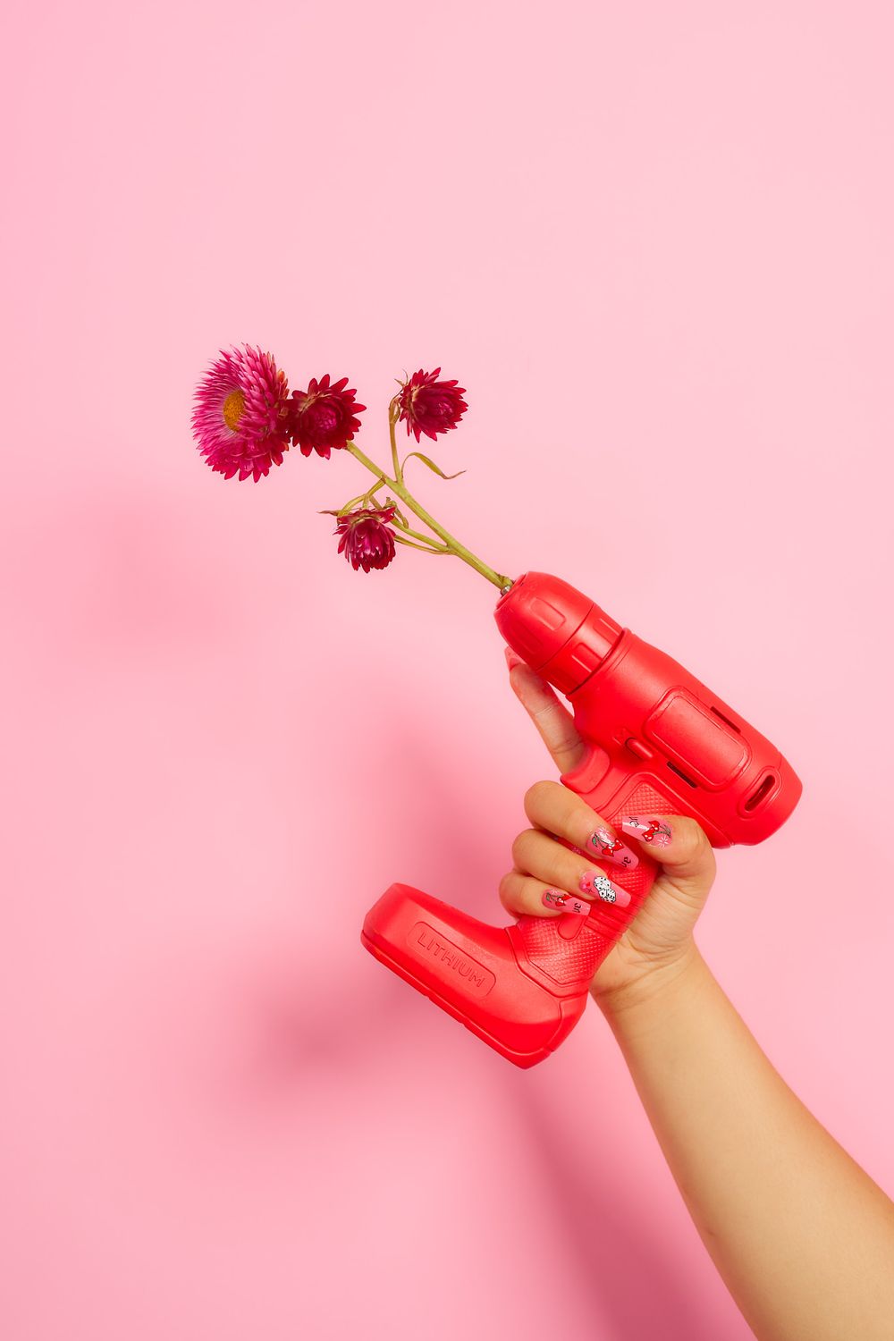 A hand holding a red power drill with pink daisy flowers emerging from the barrel on a pink background.