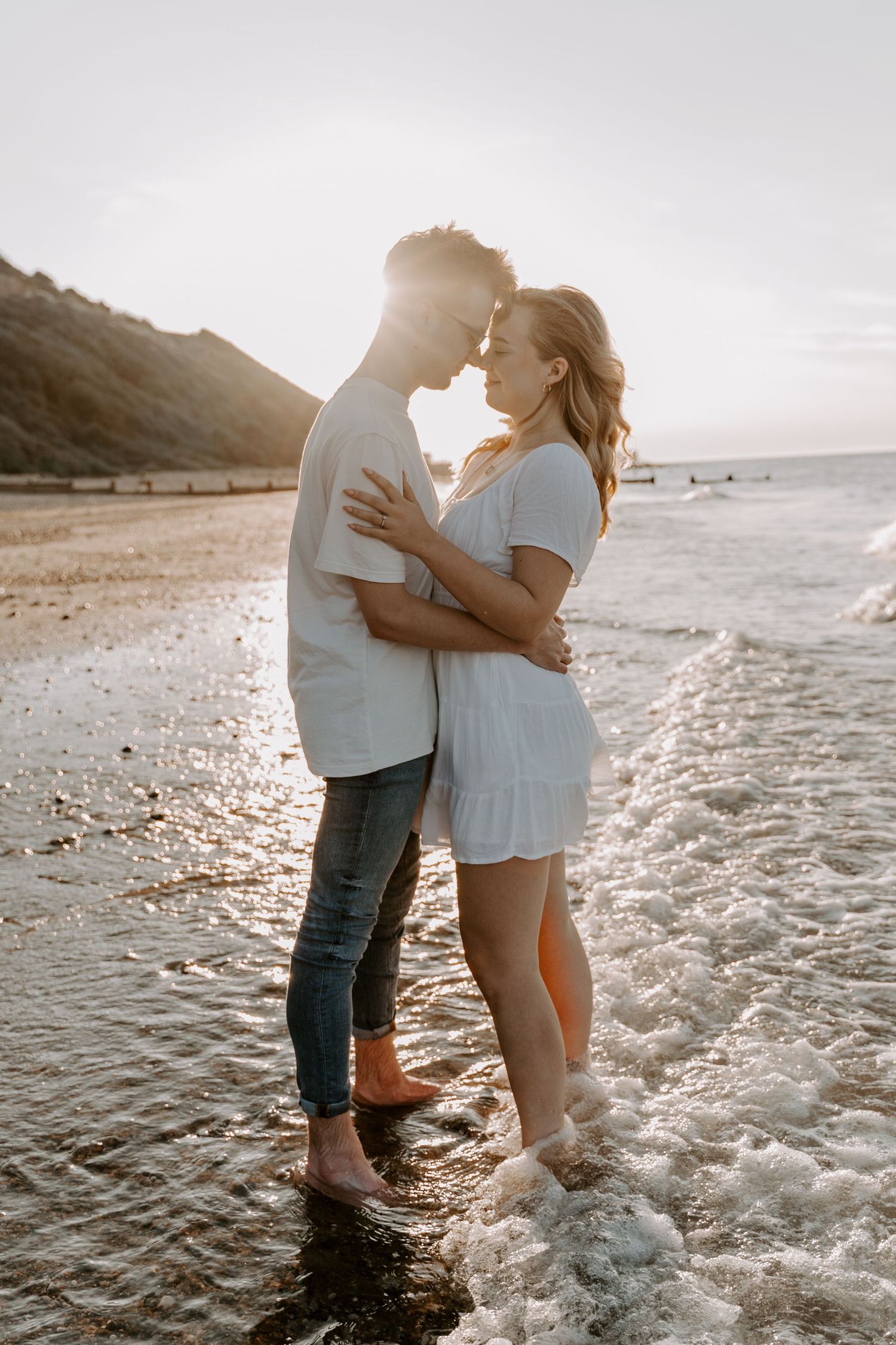 a couple are stood in the sea edge with water washing over their feet. they are touching foreheads and the sun is setting behind them.