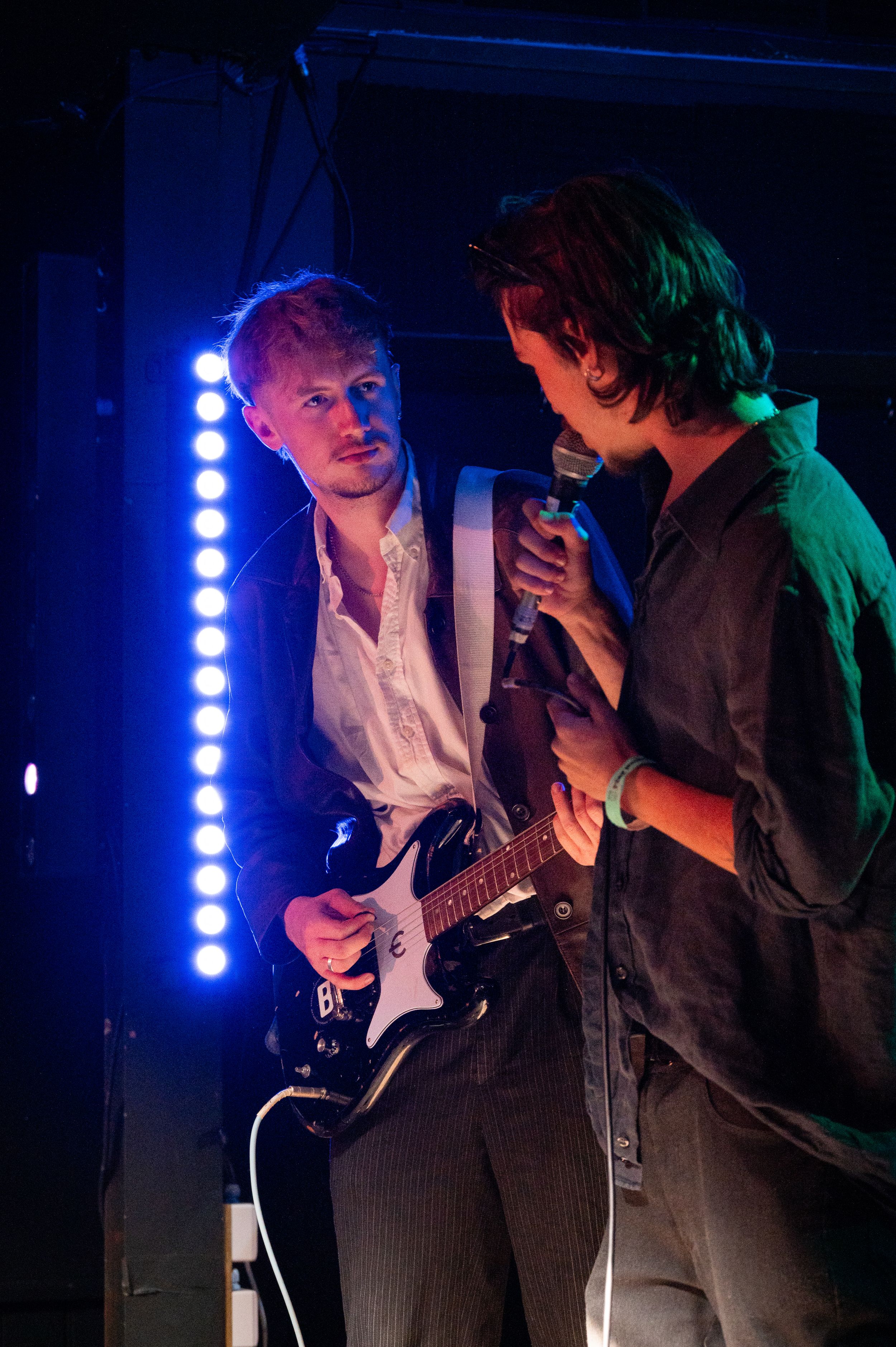 Two musicians perform on stage with electric guitar under blue concert lighting.