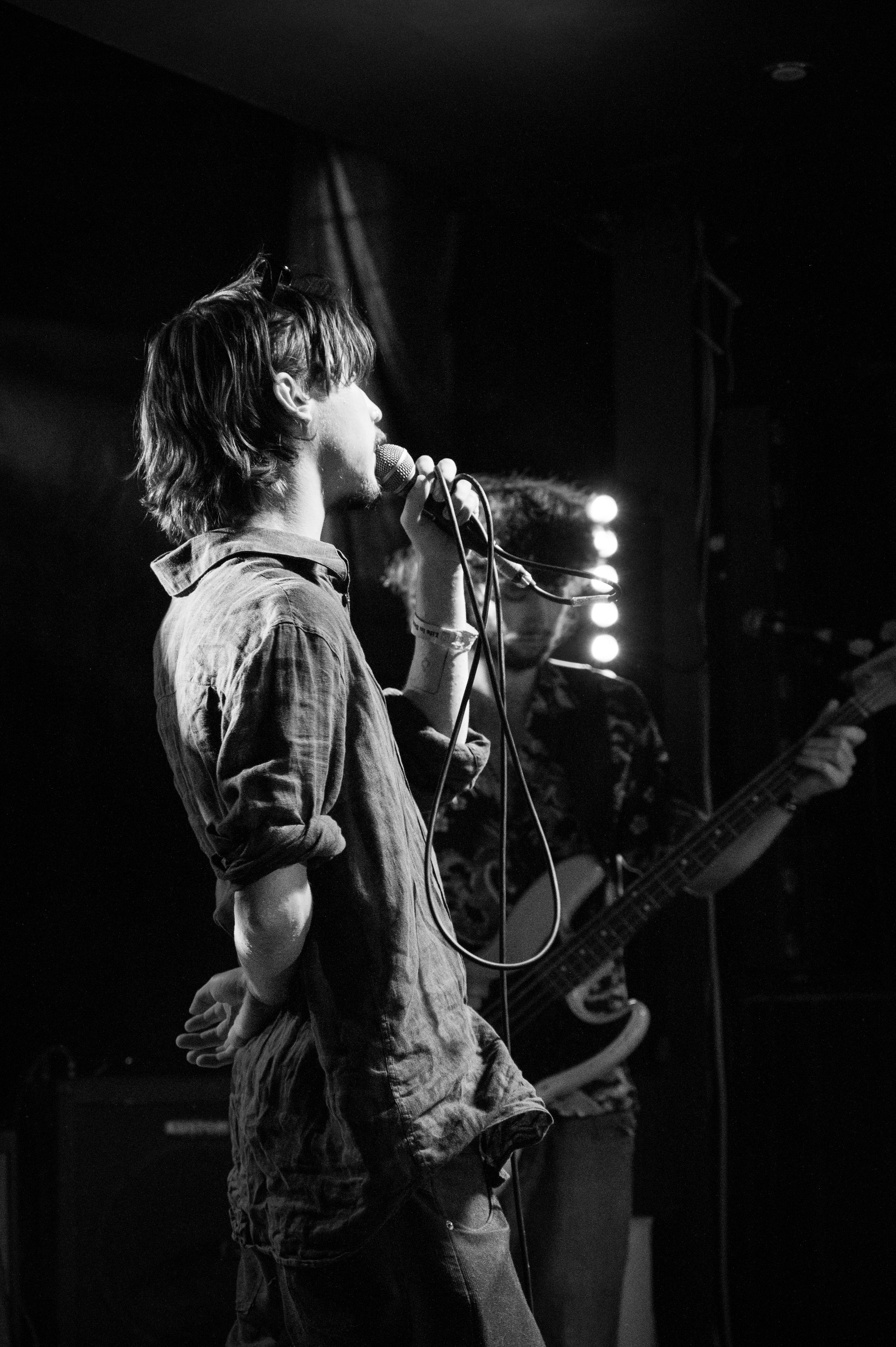 Black and white concert photo of vocalist singing into microphone on dimly lit stage.
