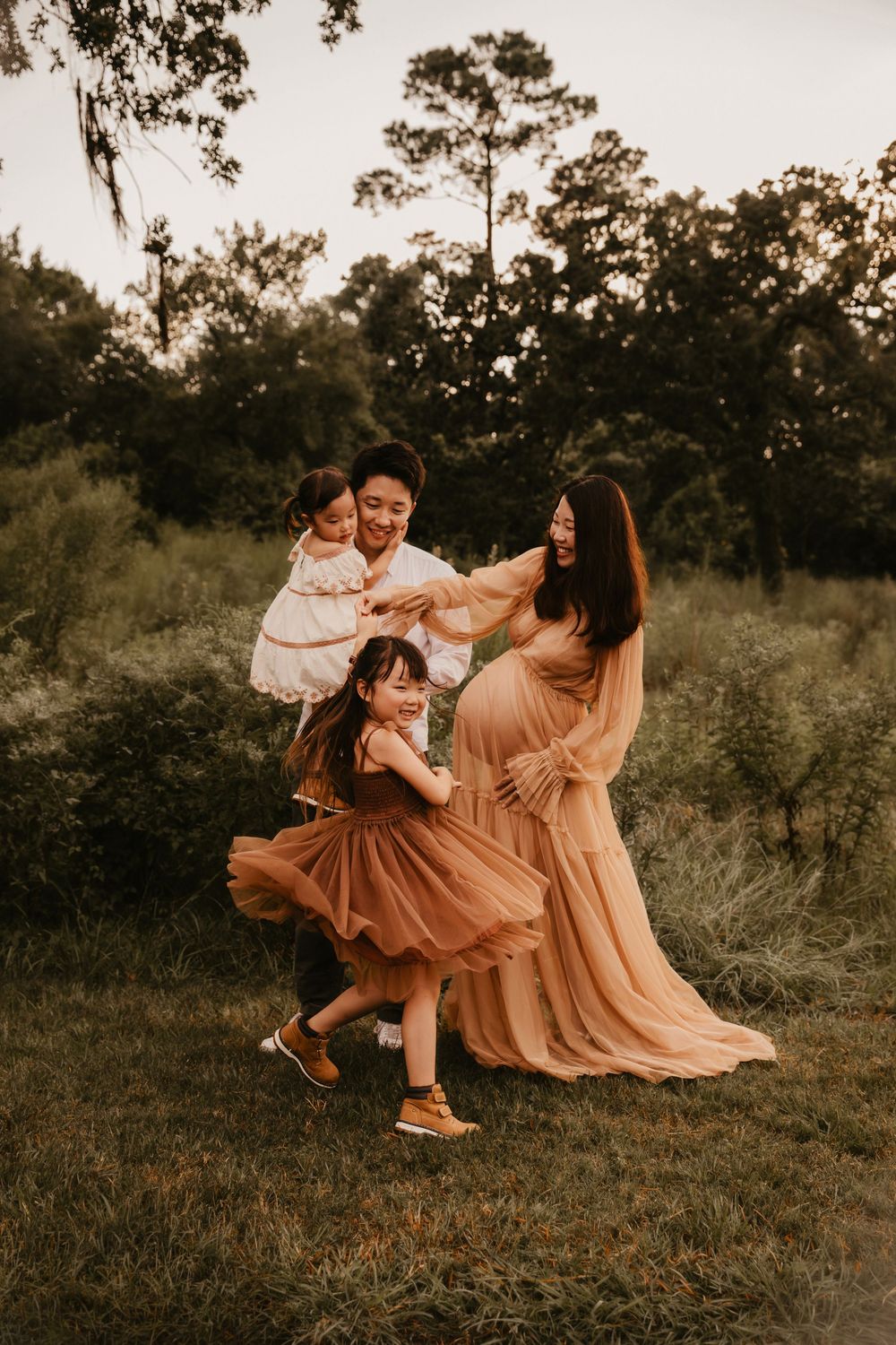 Pregnant Asian woman in flowing golden dress poses with family in lush green outdoor setting at golden hour.
