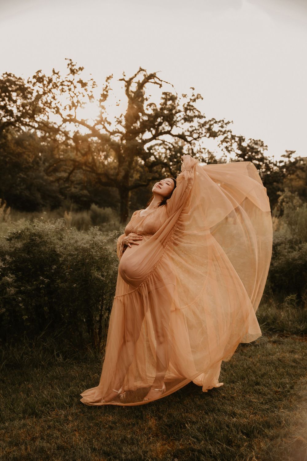 Pregnant woman in flowing peach gown poses outdoors at golden hour, fabric billowing in wind near lush green trees.