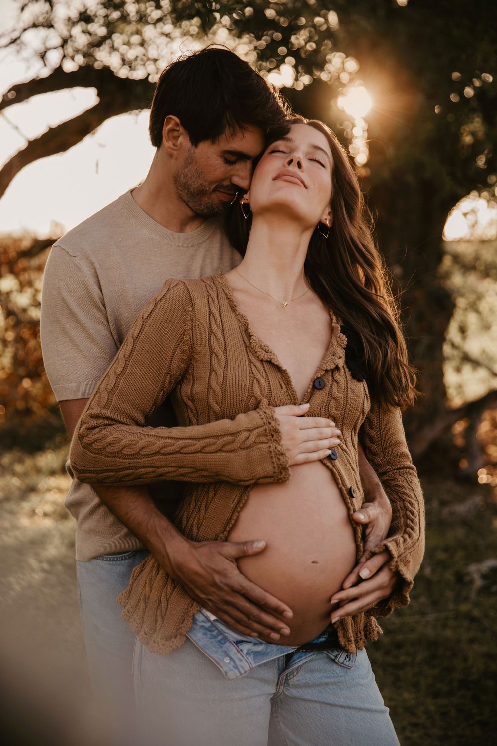 A couple shares a tender maternity moment outdoors at golden hour, with man embracing pregnant woman from behind.