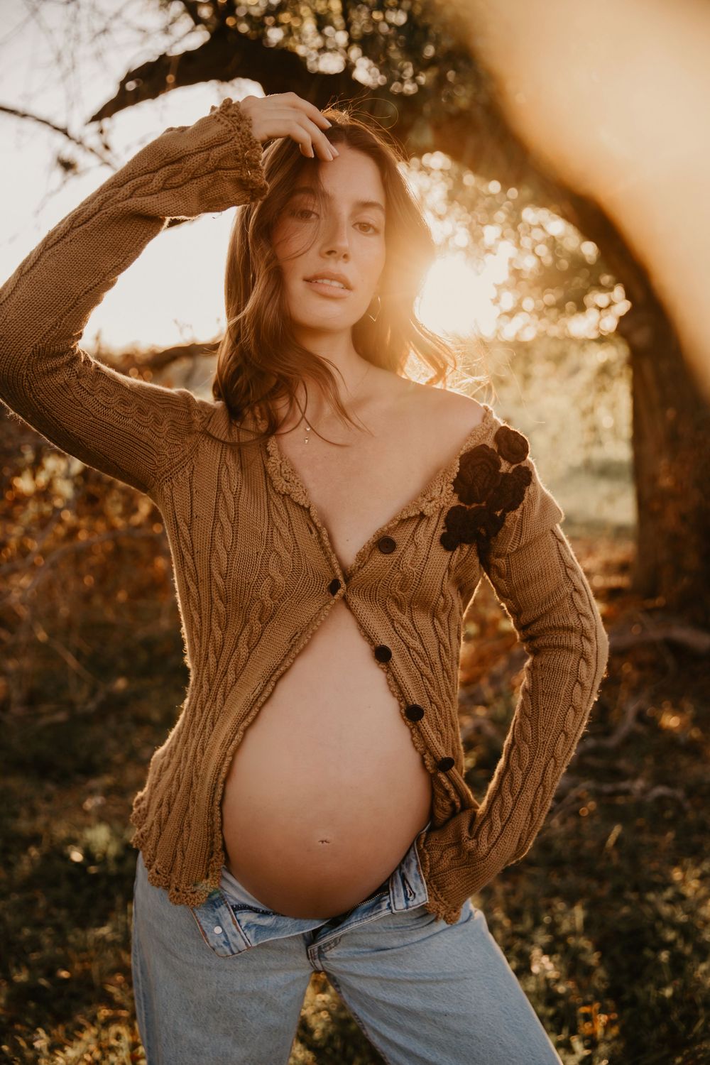 Pregnant woman in open cable-knit cardigan and jeans posing outdoors at golden hour sunset.