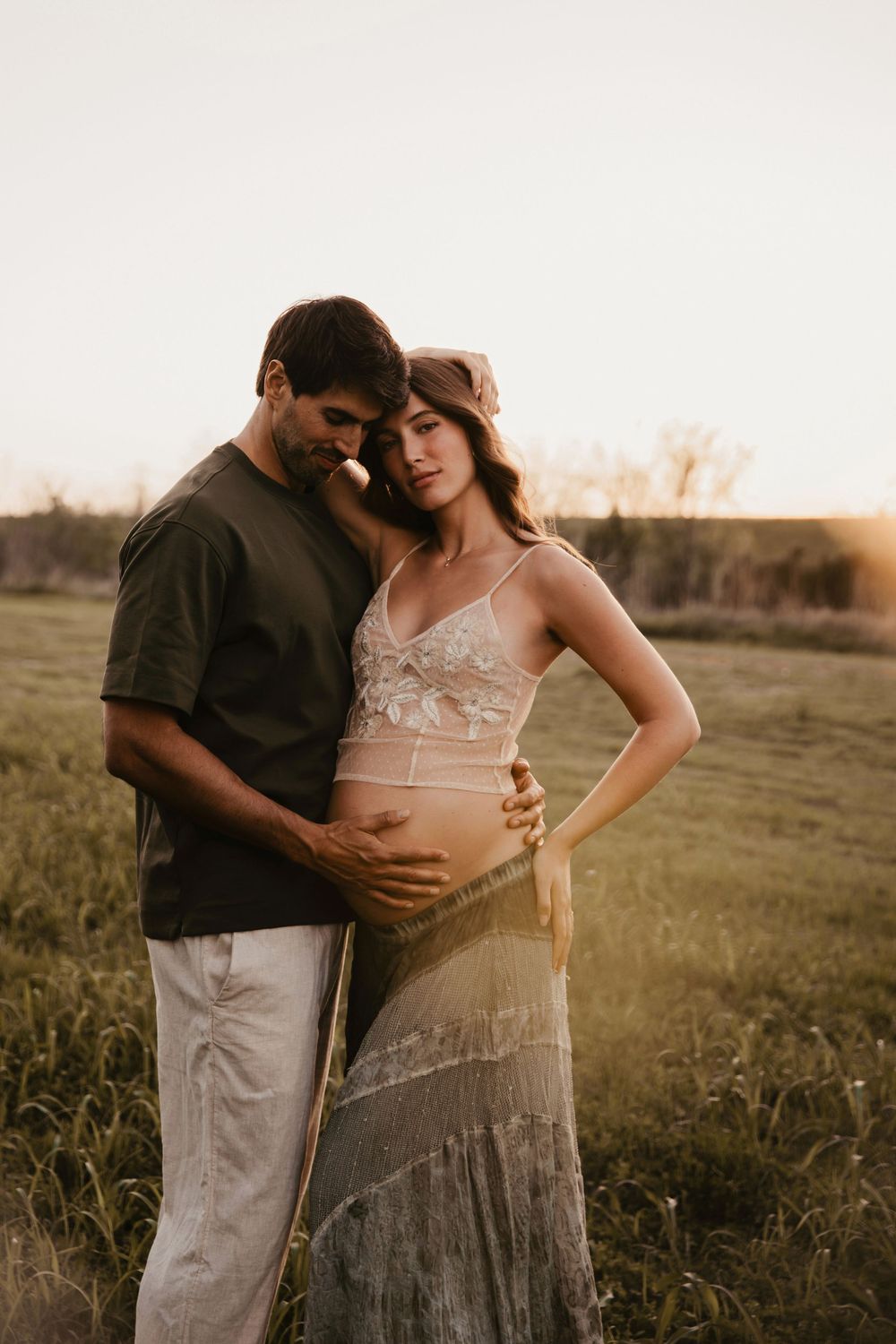 Couple embracing at golden hour in field, woman in lace maternity dress showing baby bump.