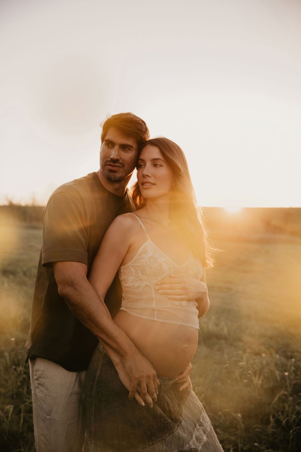 A couple poses at golden hour in a field, the pregnant woman in lace while her partner embraces her from behind.