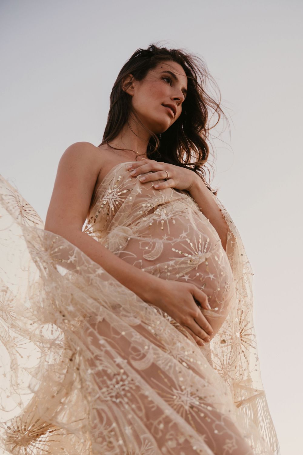 Pregnant woman in flowing star-embroidered nude tulle gown gazes softly into distance.