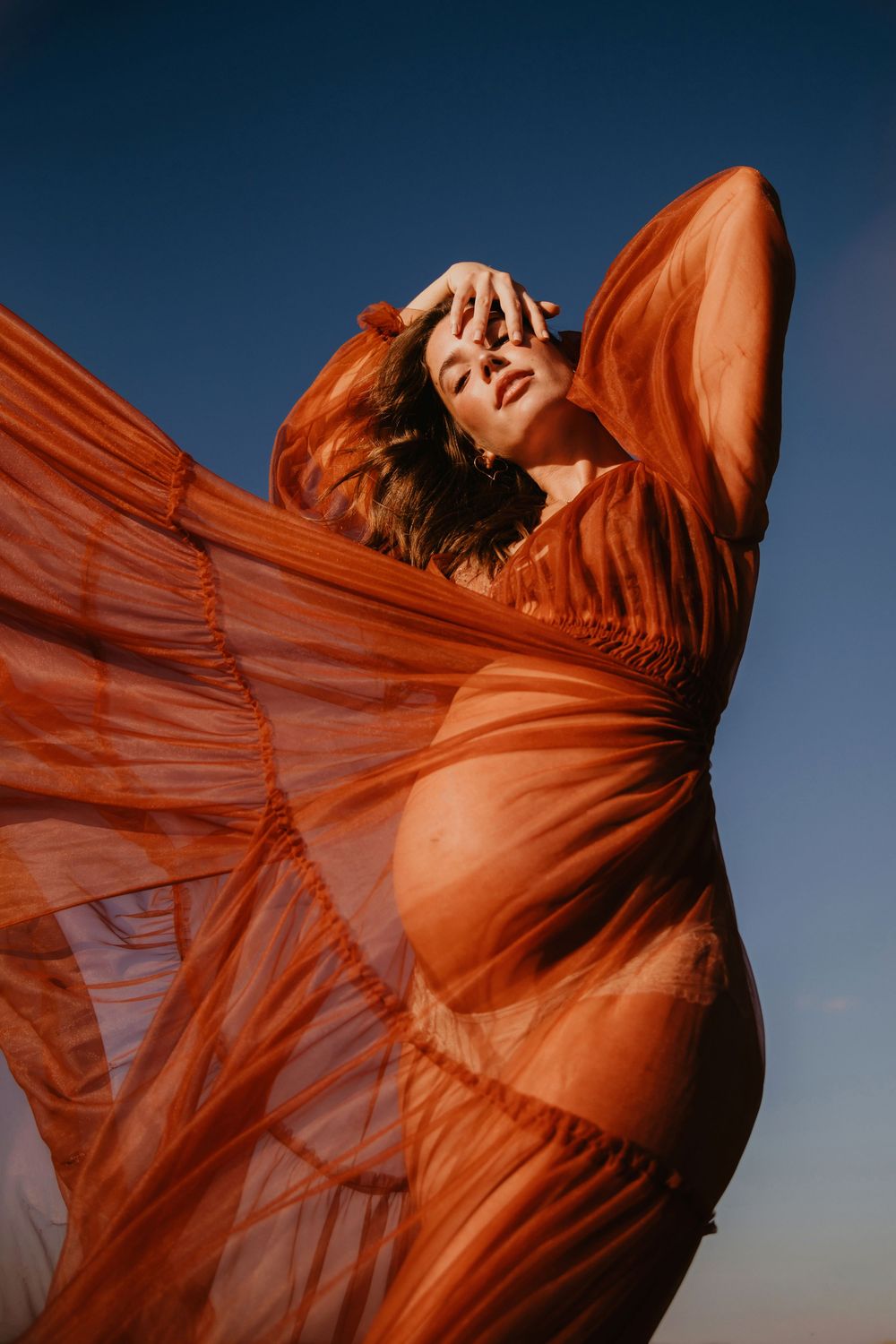 Pregnant woman in flowing rust-orange sheer dress poses dramatically against blue sky.