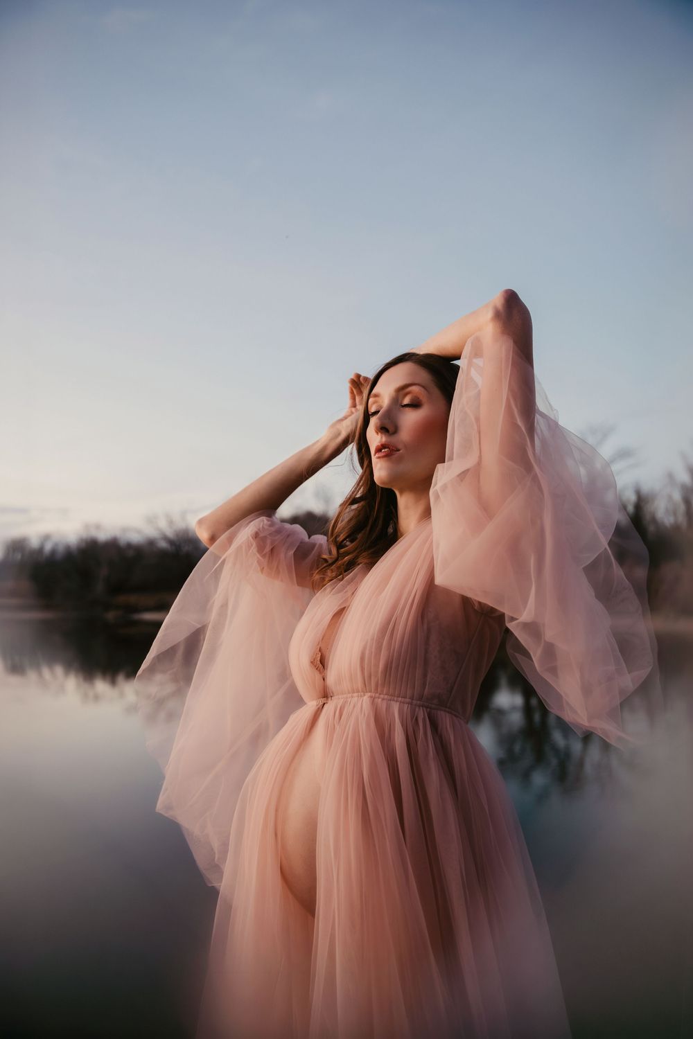 Ethereal figure in flowing pink tulle gown poses gracefully against misty lake at sunset creating dreamy atmosphere.