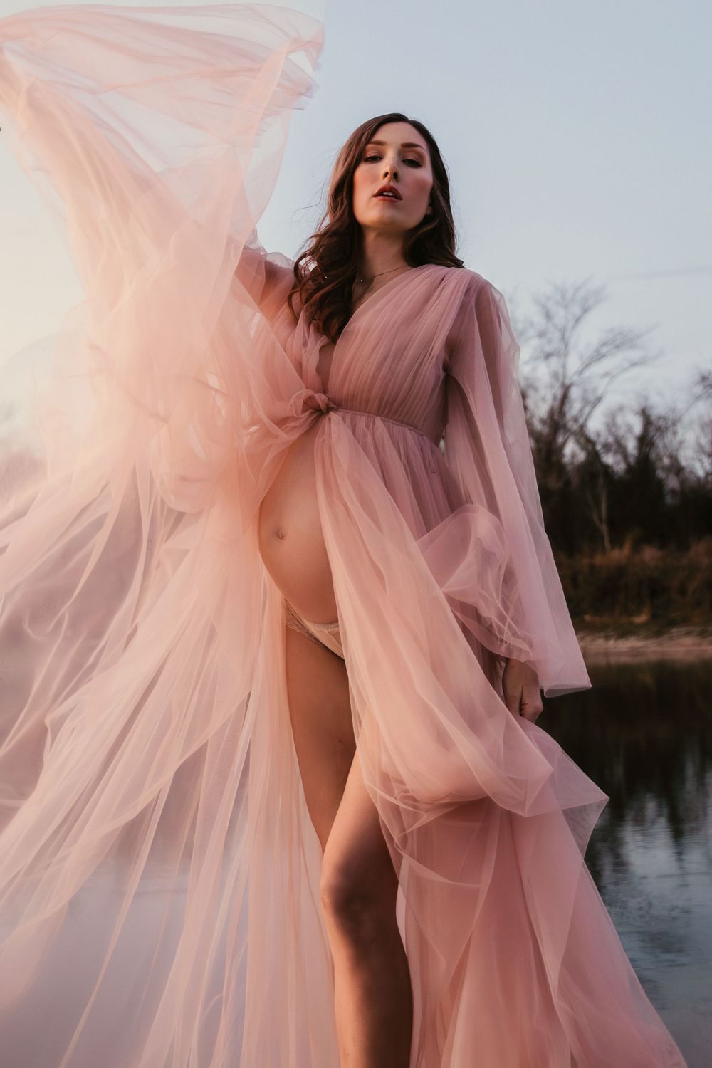 Flowing pink sheer maternity gown billows dramatically by a lake during sunset photoshoot.