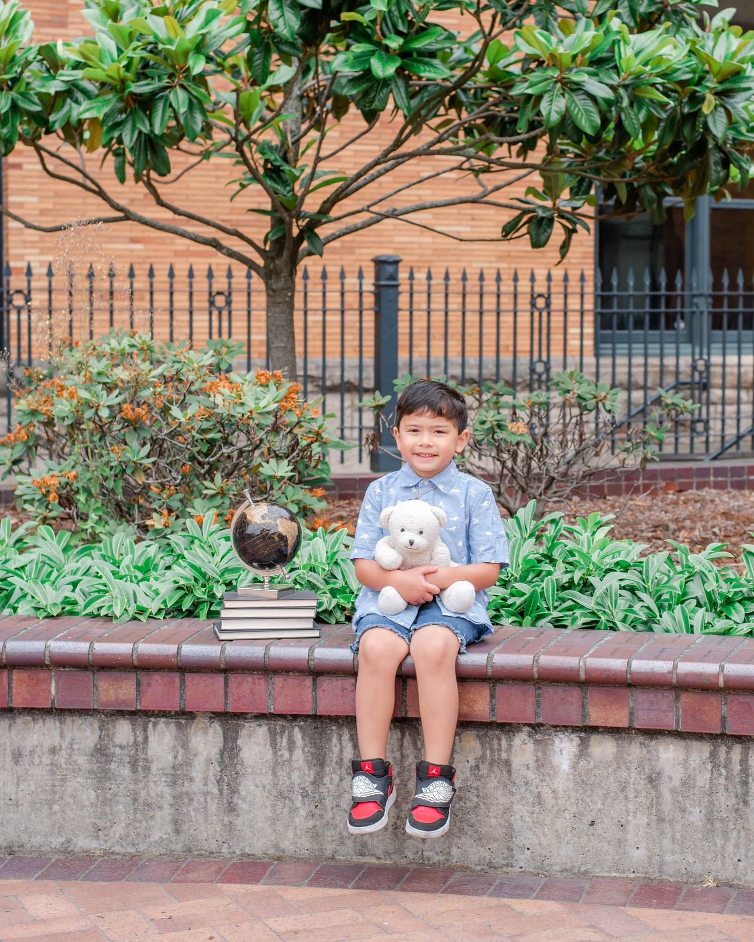Child in blue shirt sits on brick ledge with stuffed animal toy in garden setting.