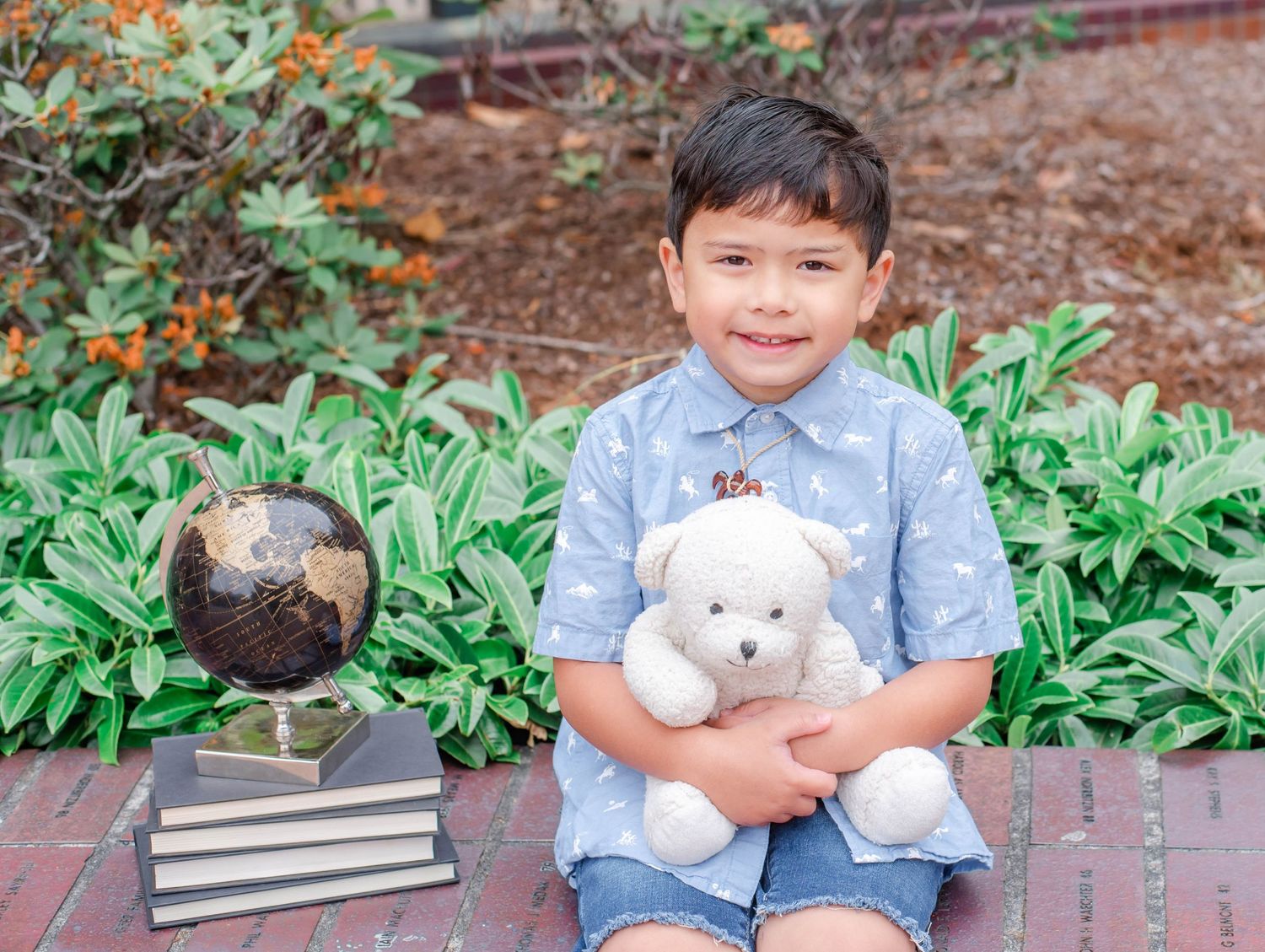 Child with stuffed animal poses on brick wall near garden plants and foliage.