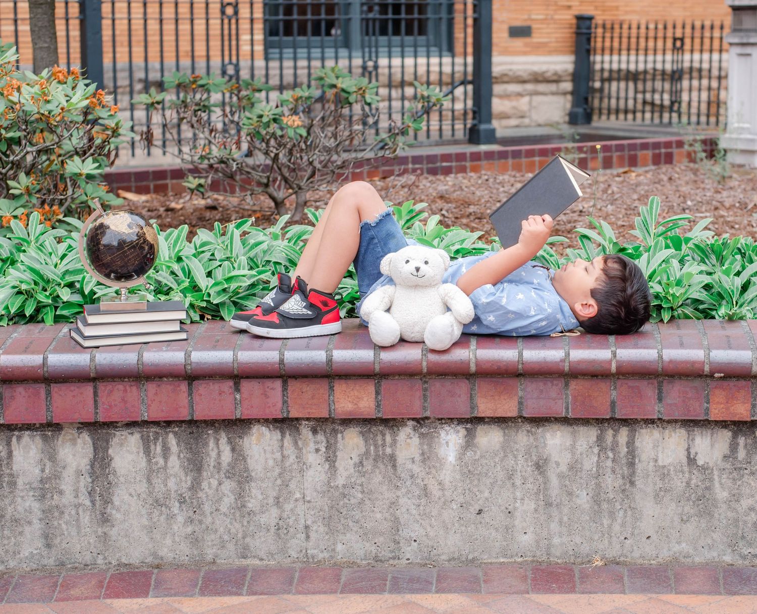 Child relaxes on brick wall with stuffed bear near garden plants and iron fence.