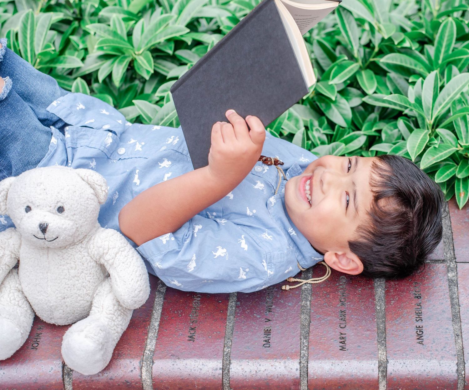 Child rests on brick ledge with plush teddy bear surrounded by garden foliage.