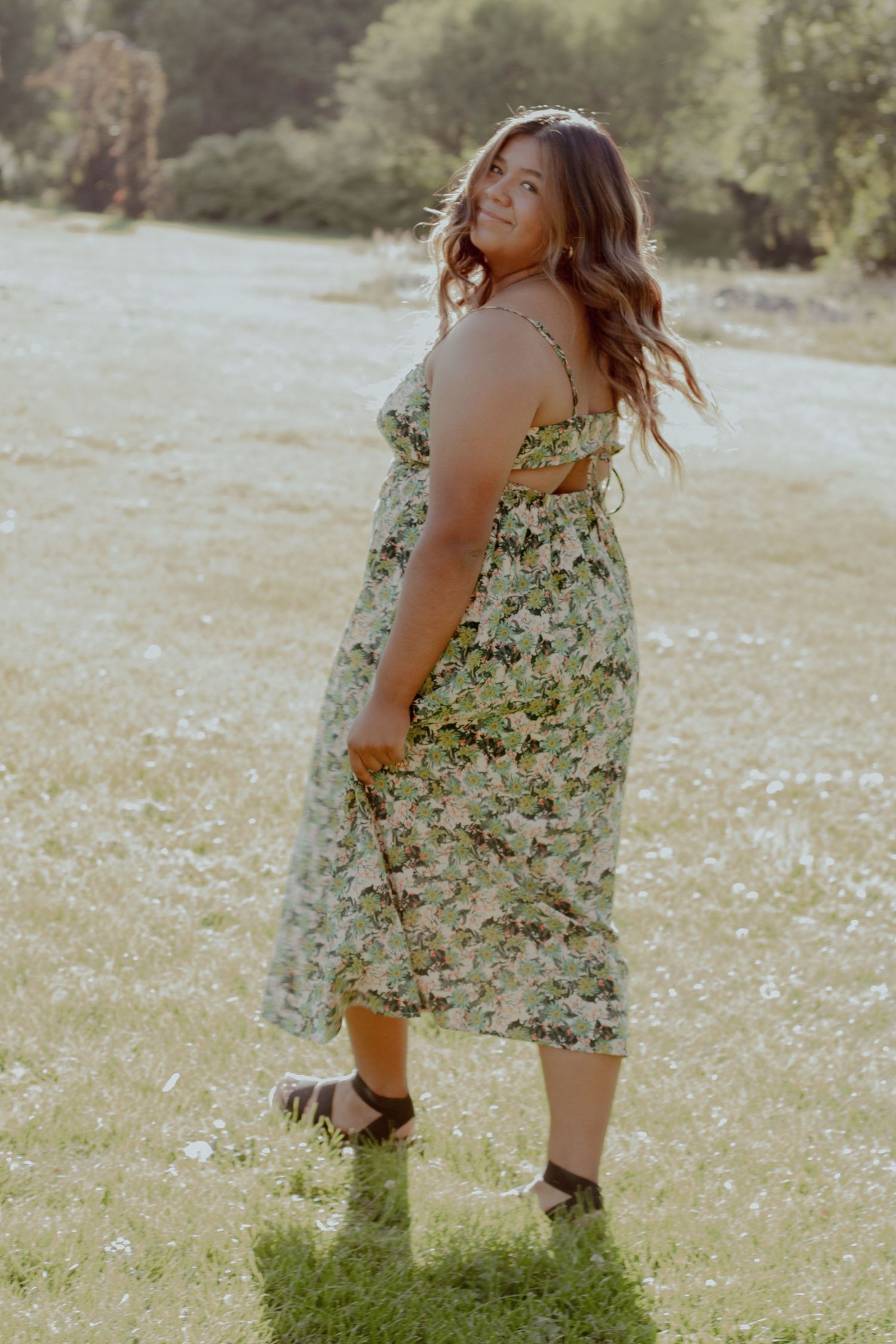 A person in a floral sundress poses playfully in a sunlit meadow filled with wildflowers during golden hour.