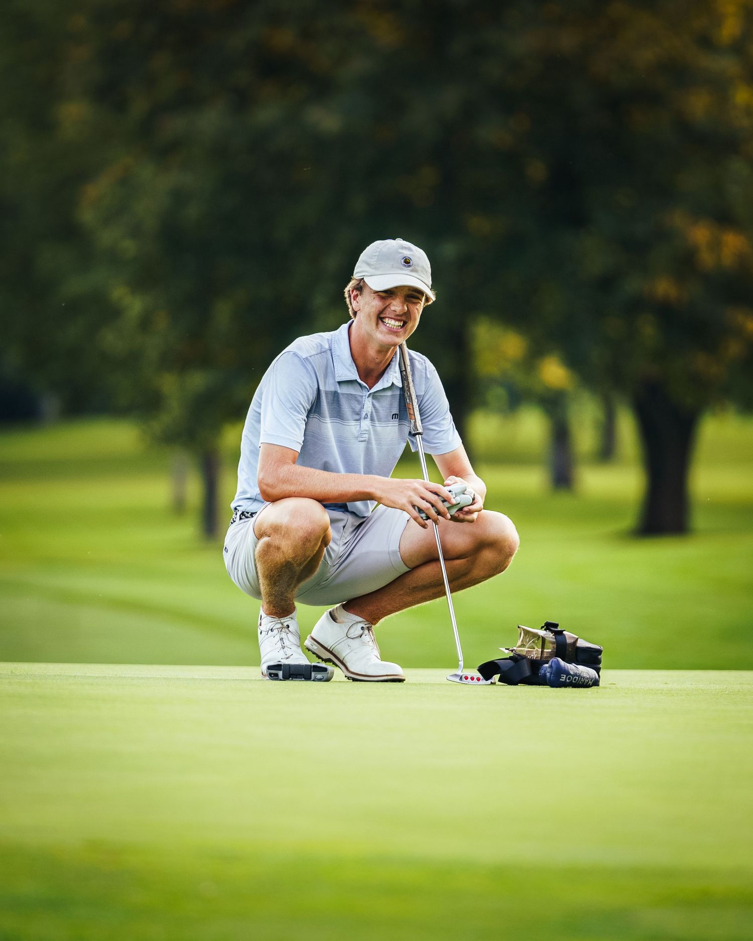 Golfer crouching on green course studying putting line with white cap and polo shirt in scenic setting.