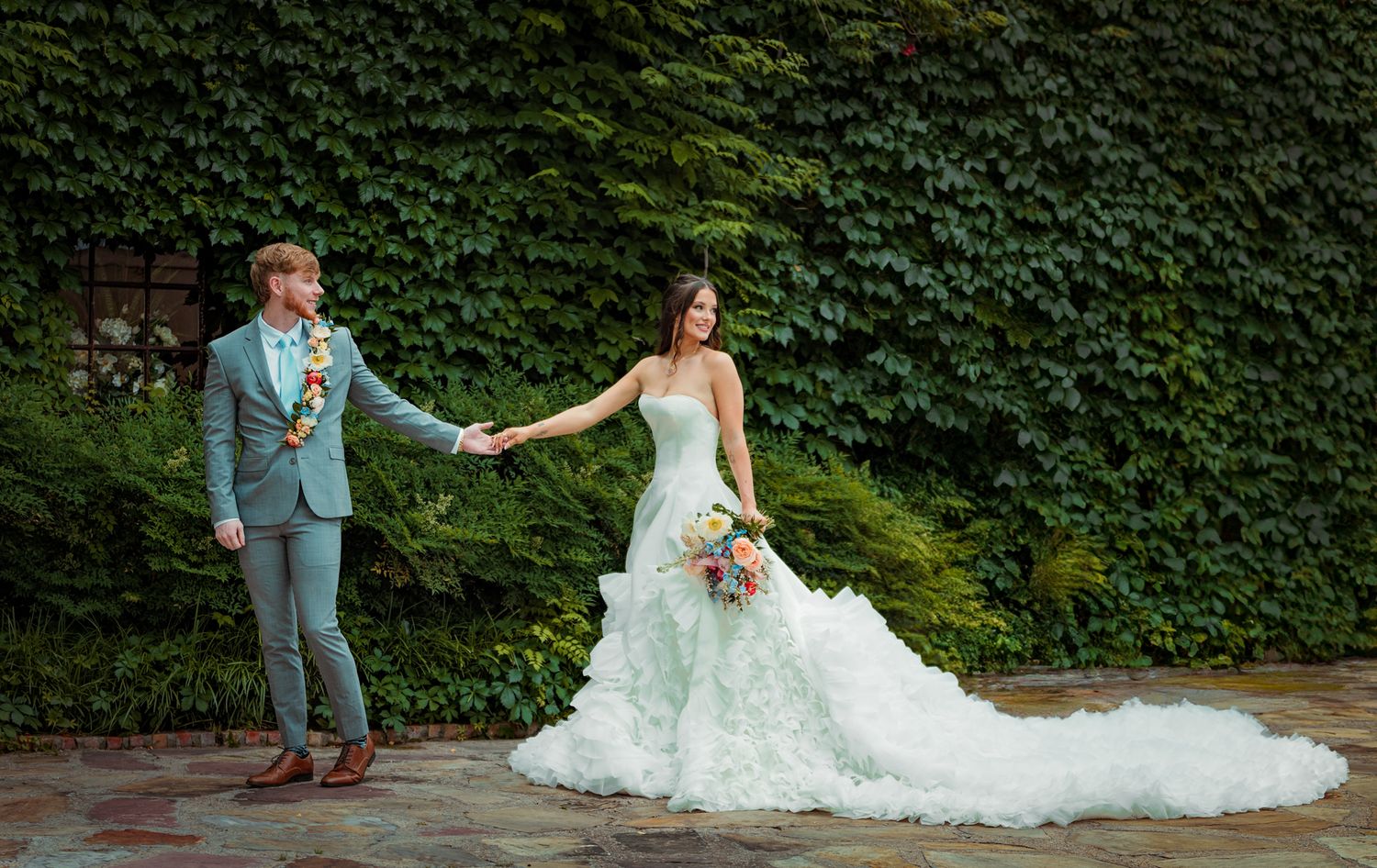 Newlyweds holding hands against ivy-covered wall in formal wedding attire.