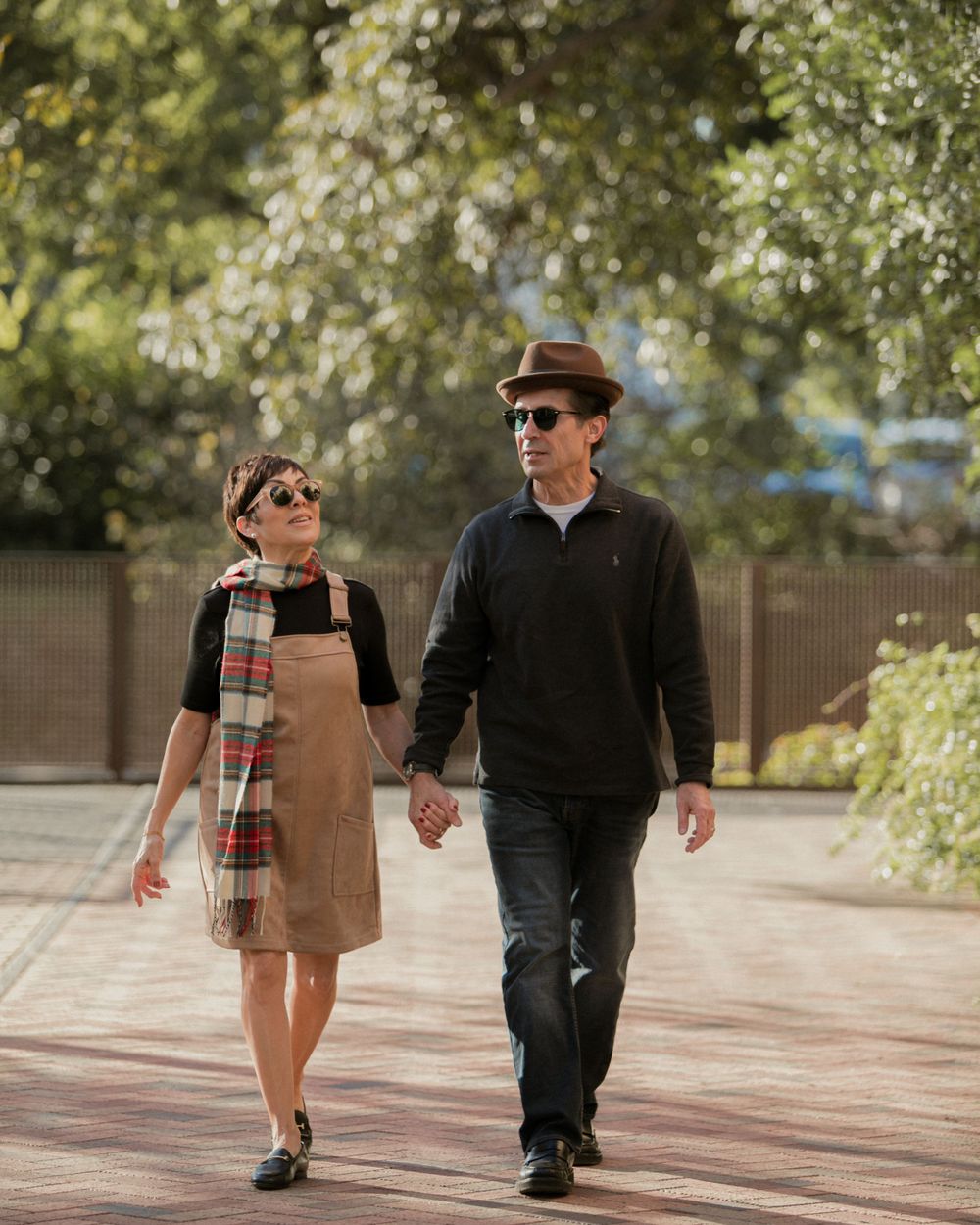 A couple walks hand in hand on a brick path with autumn trees in the background.