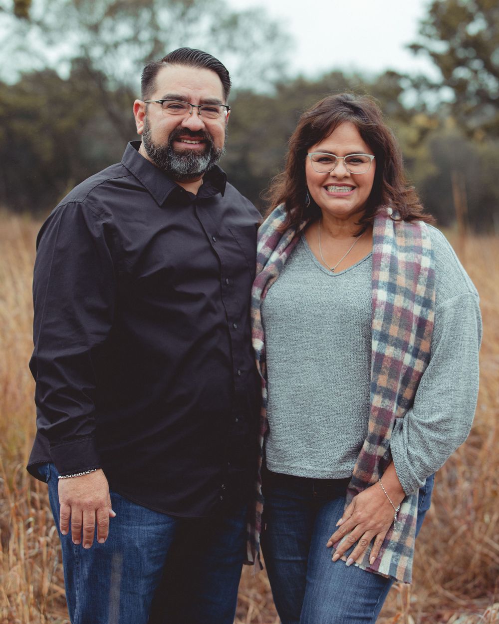Couple in plaid and casual wear smiling together in autumn field setting.