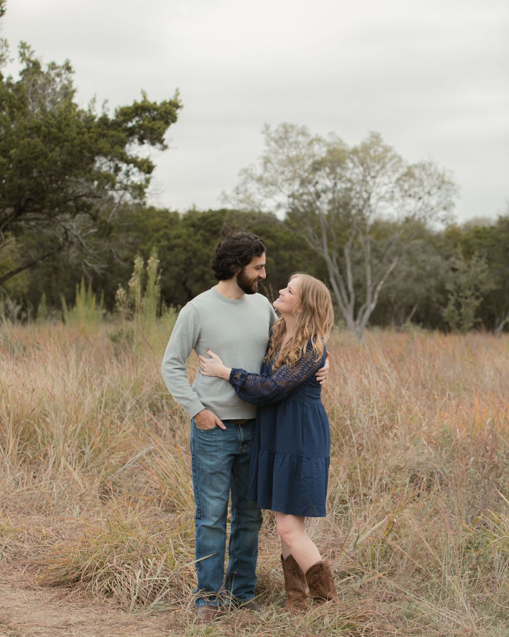 Couple in casual attire embracing in tall grass field during overcast day.