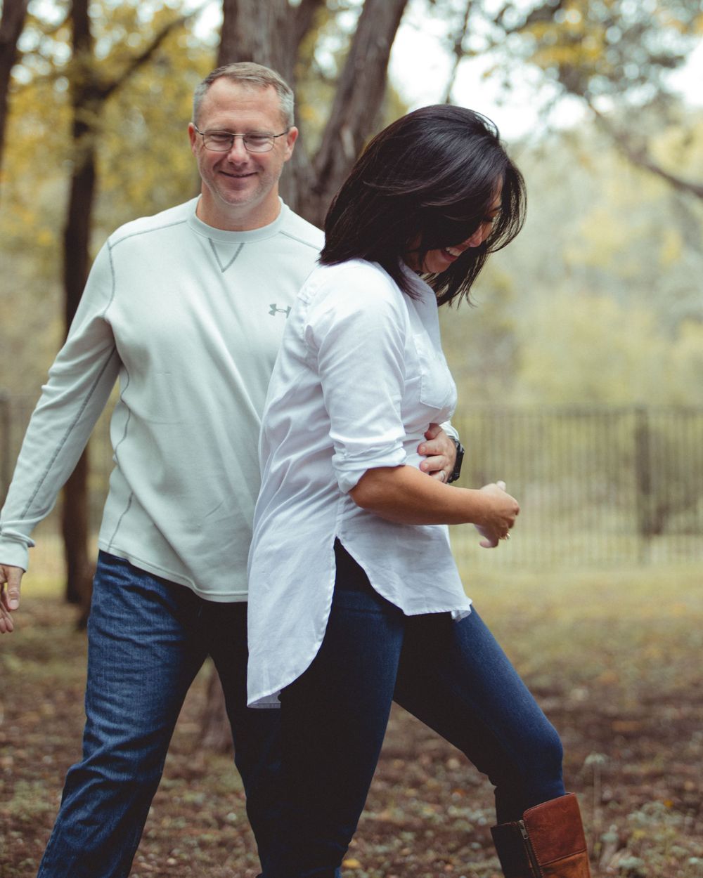 Playful couple in white tops and jeans posing together in park setting.