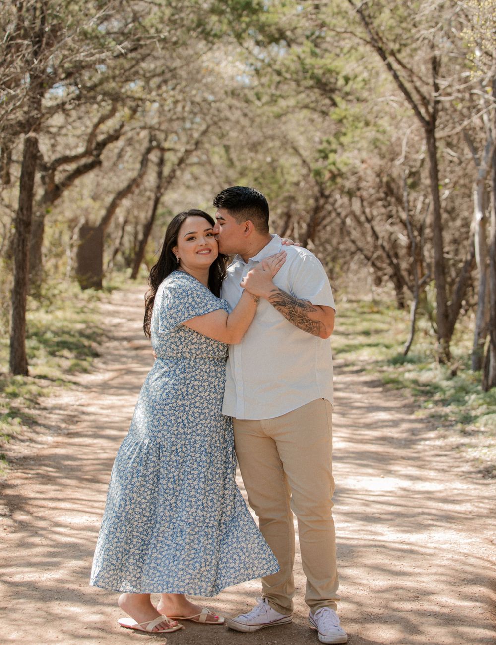 A couple shares tender moments on a sunlit dirt path surrounded by trees.