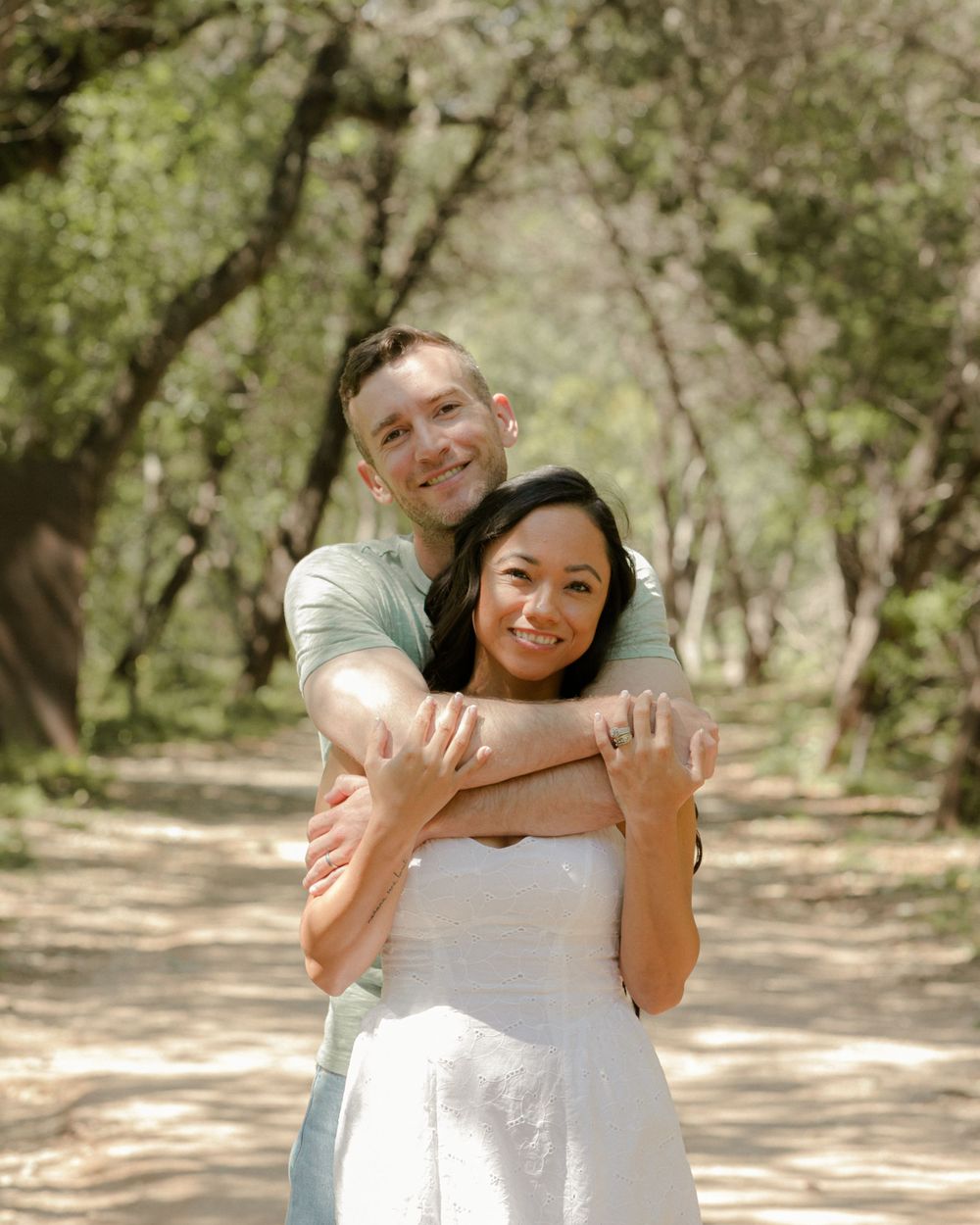 A couple embraces joyfully in a sun-dappled outdoor setting with natural greenery.