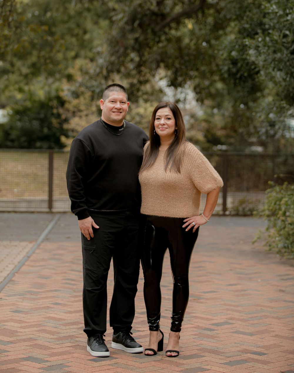 Couple in black and beige outfits posing together on brick pavement in outdoor setting.