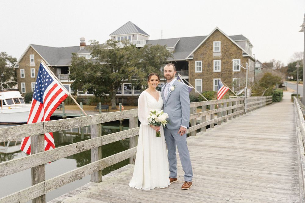 A couple walks along a wooden boardwalk with American flags and coastal houses in the background.