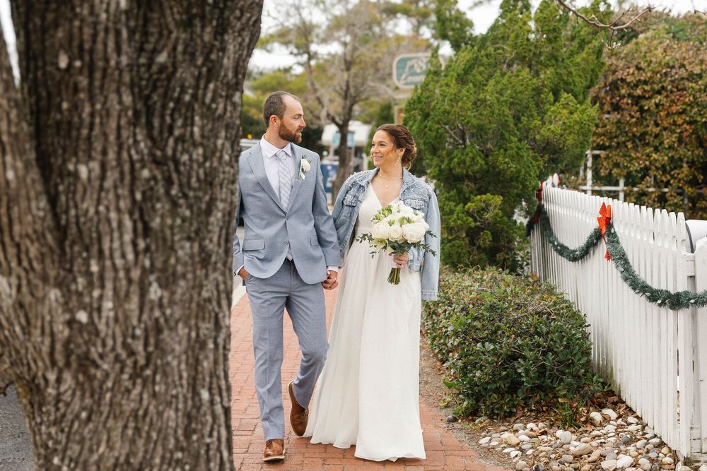 Newlyweds walking along a white picket fence path surrounded by trees.