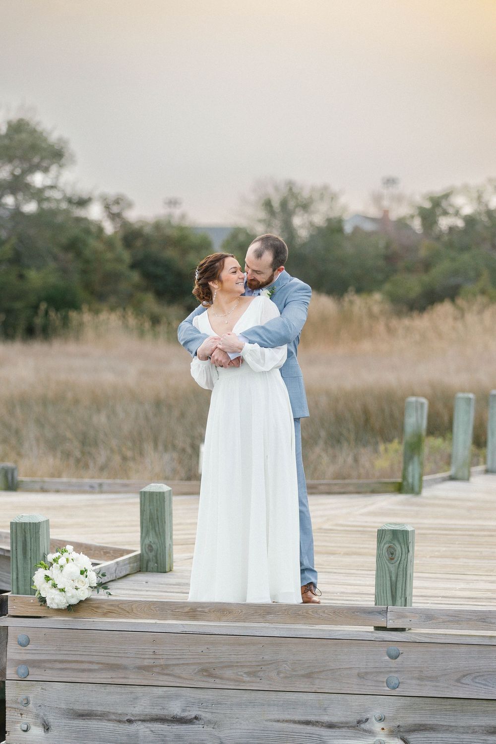 Couple embracing on wooden dock with coastal marsh grass in background.