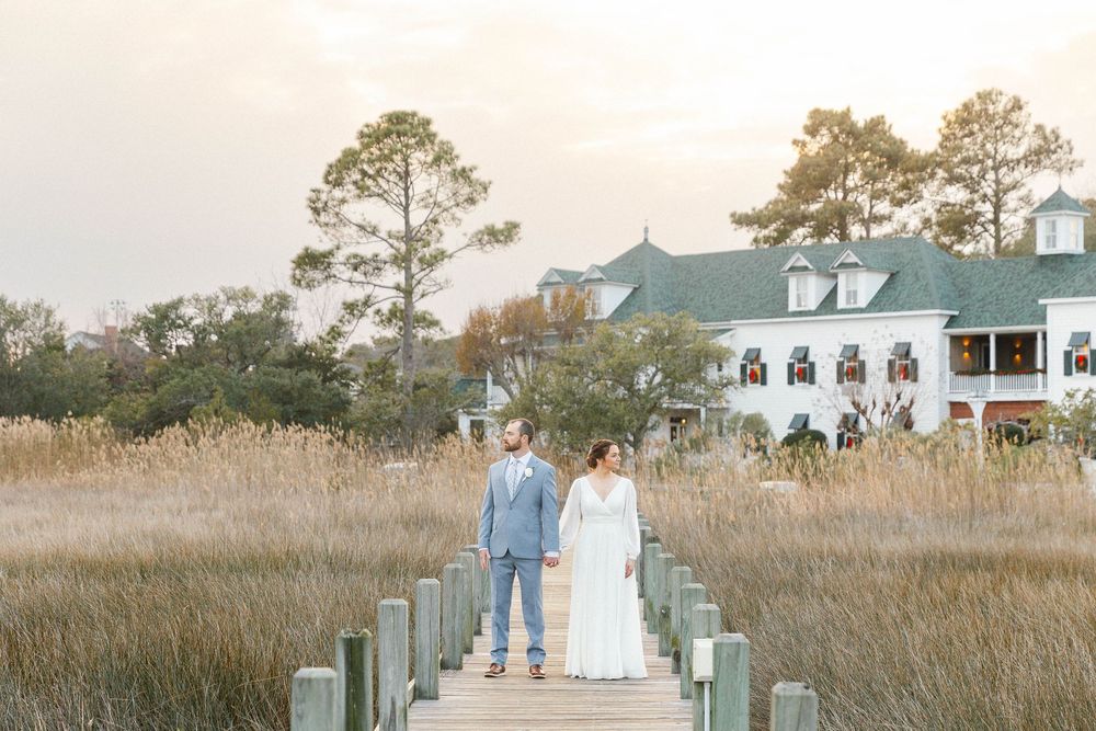 Wide view of coastal boardwalk with pine trees and marsh grass at sunset.