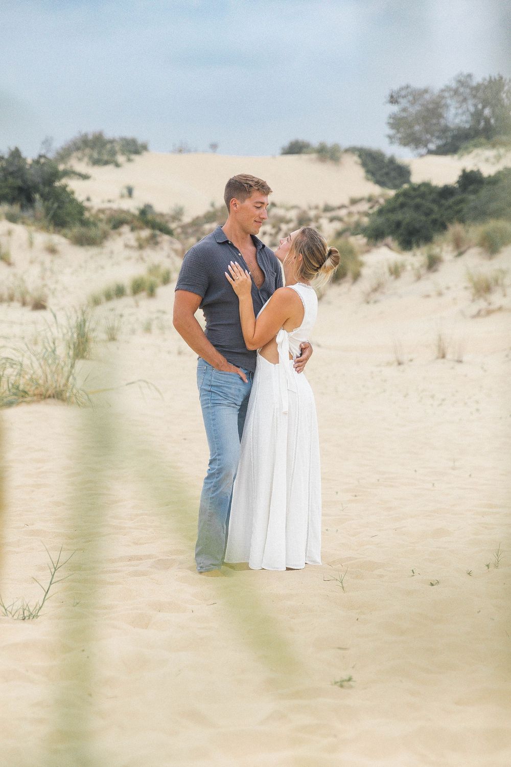 A couple shares an intimate moment on sandy beach dunes with coastal vegetation in the background.
