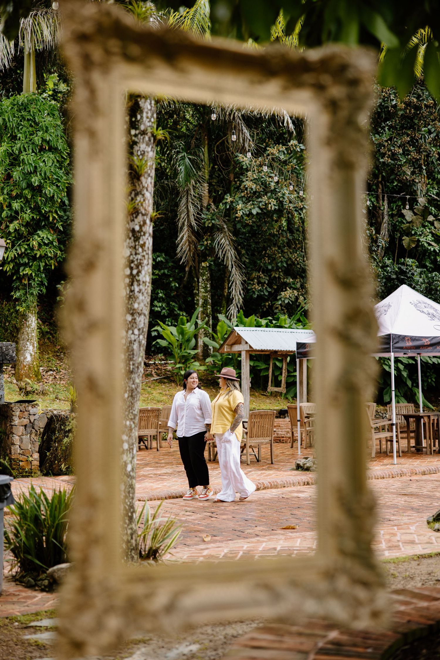 A romantic couple is framed through an ornate vintage frame in an outdoor garden setting with tents visible.