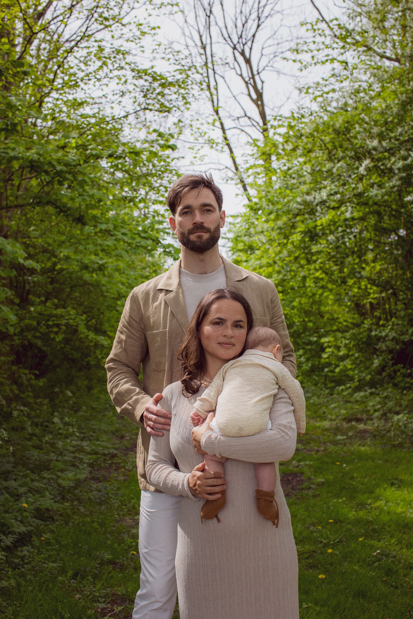 Couple in neutral-toned outfits embrace tenderly in a lush green outdoor setting.