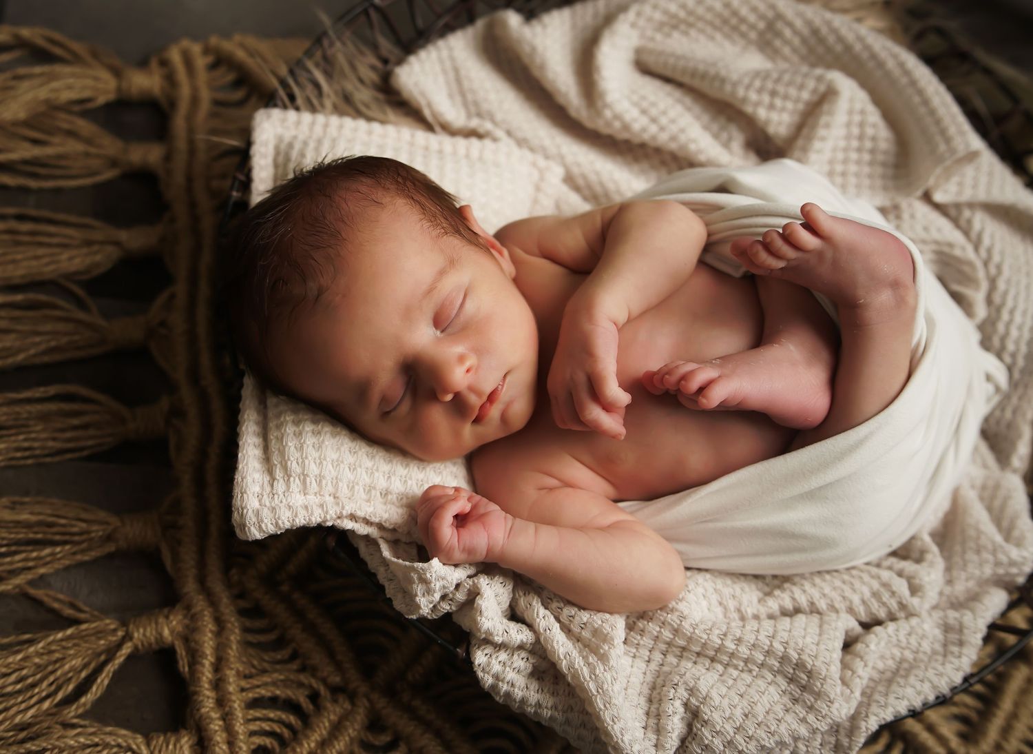 Sleeping newborn baby rests on a soft cream blanket and woven rope backdrop during a cozy newborn photography session.