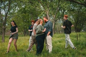 Couples and friends dancing and embracing in a lush green meadow surrounded by trees on a warm day.