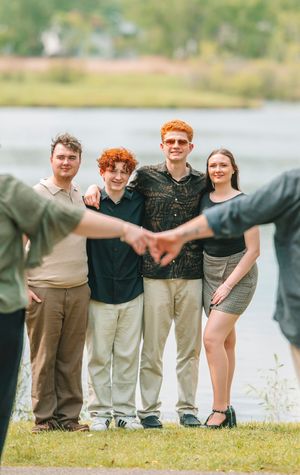 Four young adults smile together outdoors near a lake, with two people forming a heart shape in the foreground.