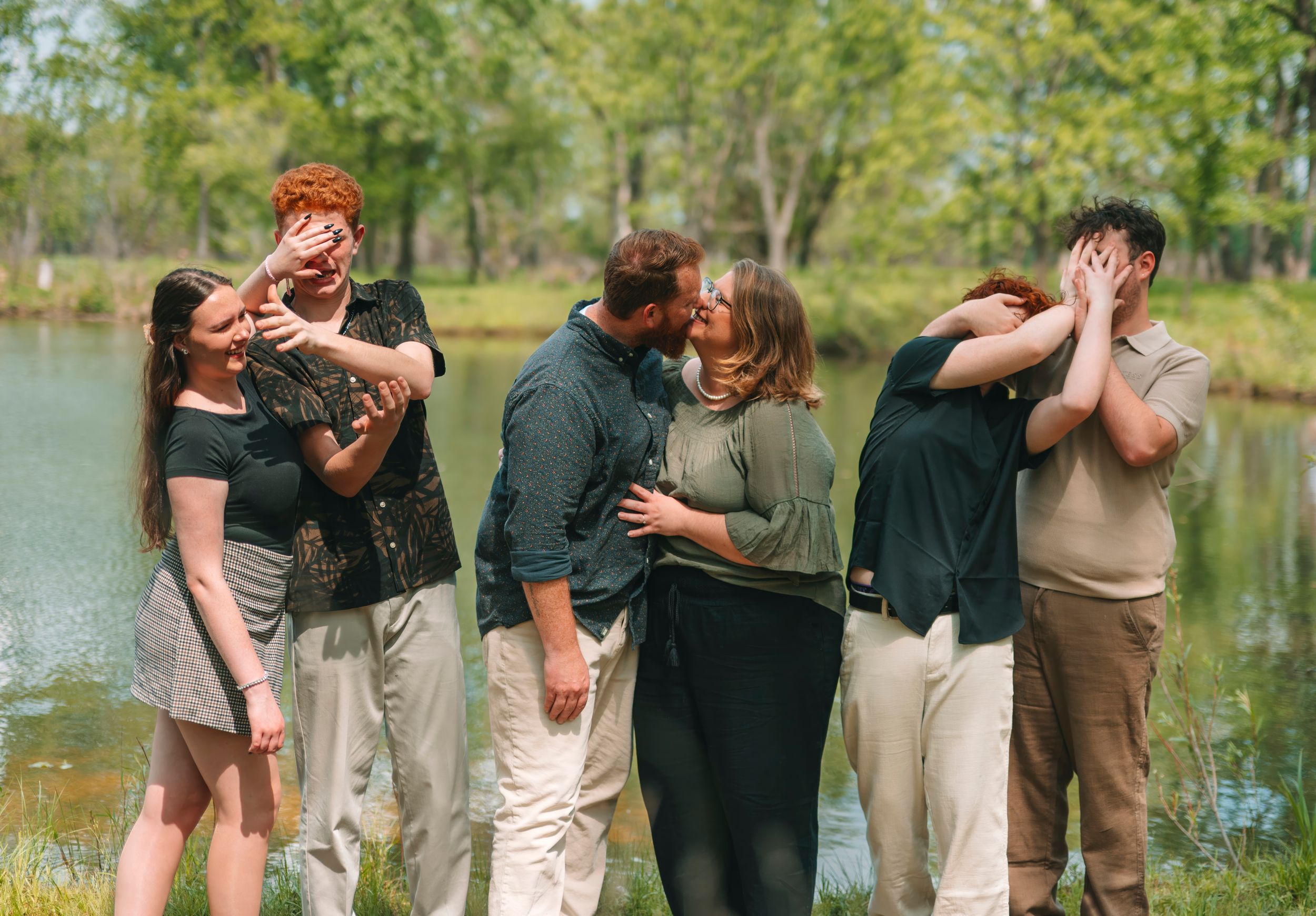 Three couples share romantic moments outdoors near a pond, surrounded by lush green trees on a sunny day.