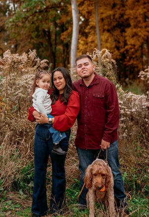 A family in red and denim outfits poses together with their golden-brown dog in a scenic autumn outdoor setting.