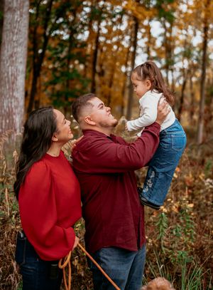 A couple in red and burgundy sweaters playfully lifts a small child in an autumn forest setting.