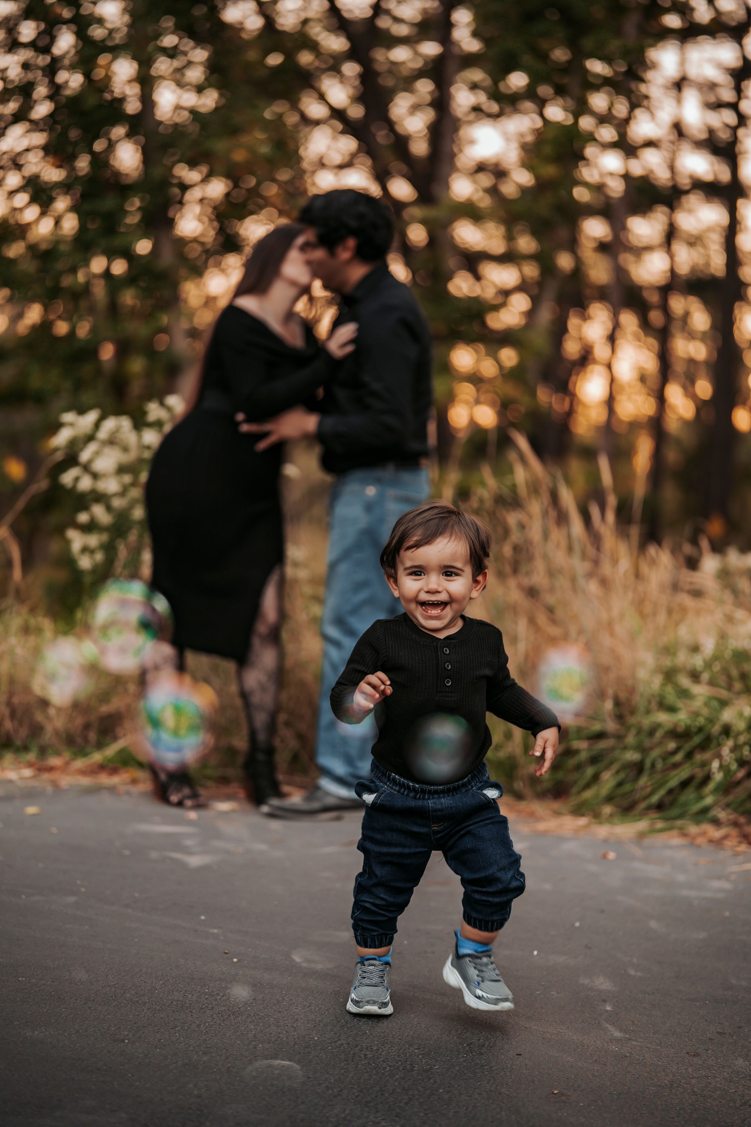 A toddler runs playfully on a path at sunset while a couple embraces in the background among autumn trees.
